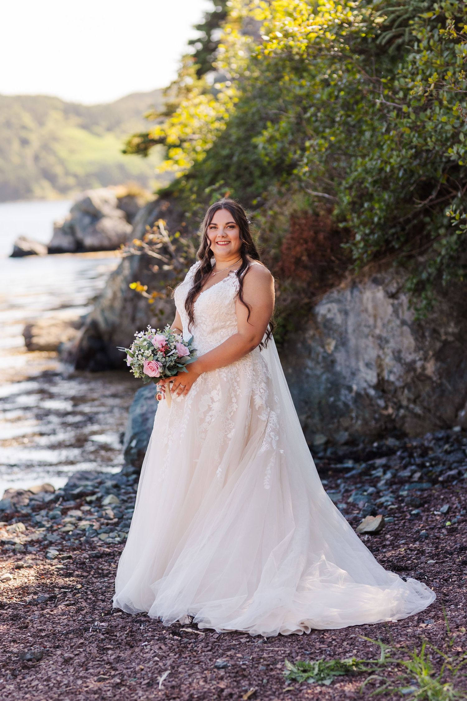 A bride in a flowing lace wedding gown poses with her bouquet on a rocky beach shoreline at sunset.