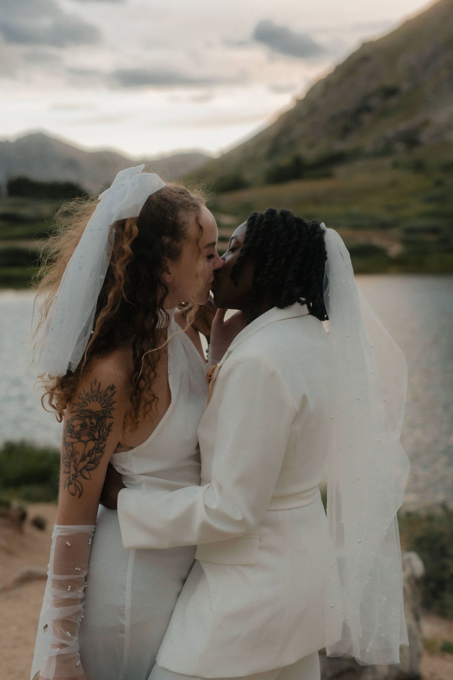 Two brides in white dresses share an intimate moment against a mountain backdrop.