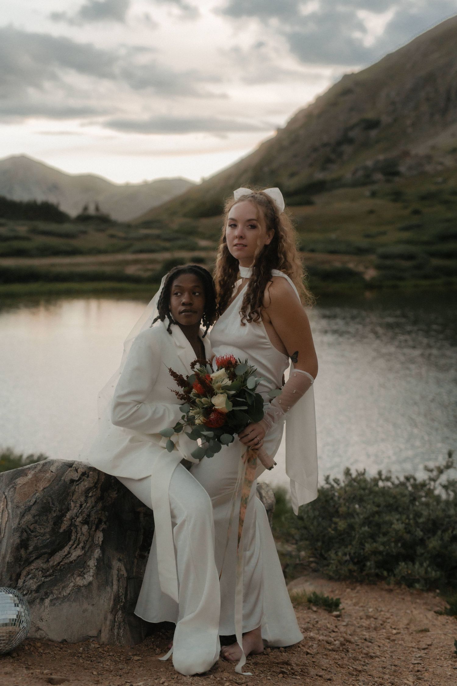 Two newlyweds in white attire pose with bouquet against mountain lake scenery at sunset.