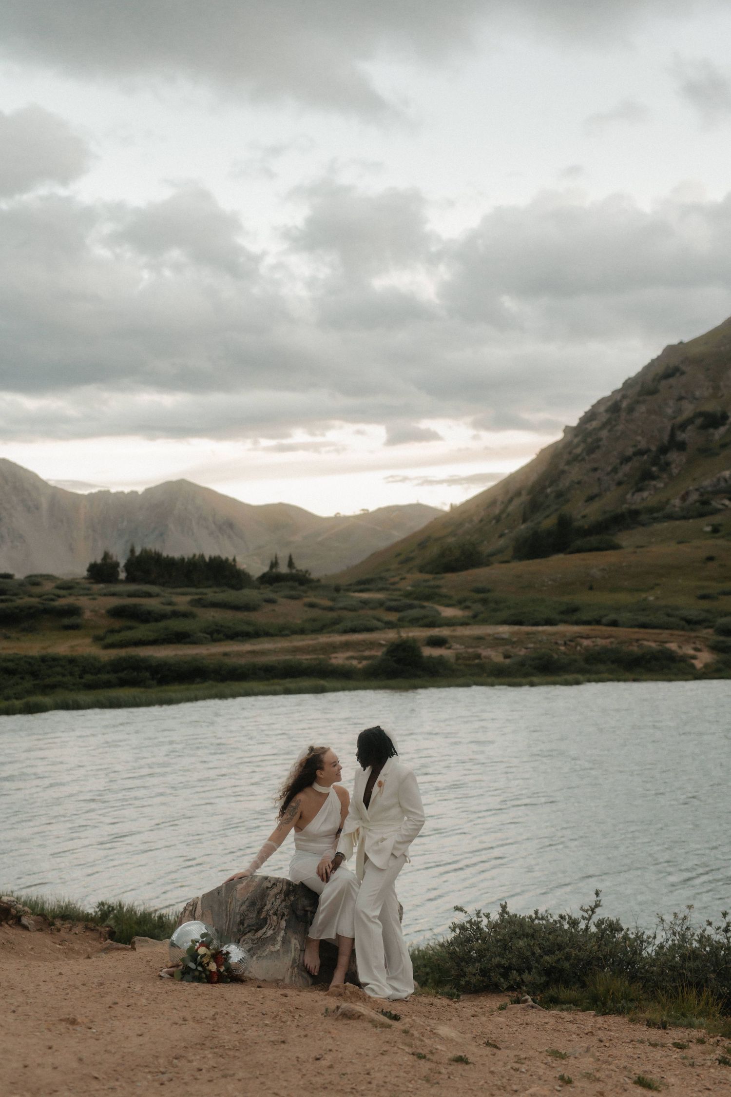Dreamy sunset landscape with a couple in white embracing by an alpine lake surrounded by mountains.