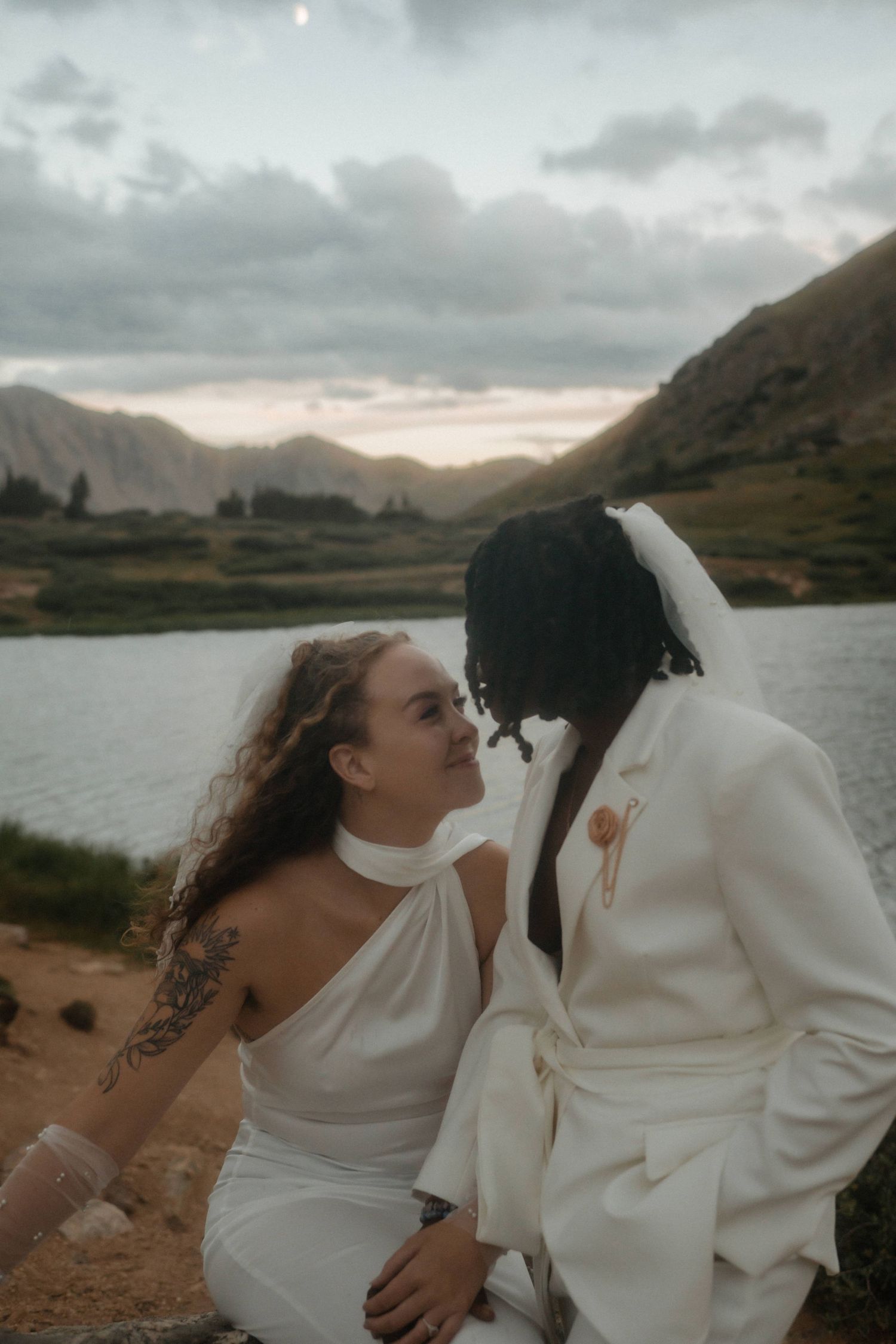 Intimate moments captured by a mountain lake with a couple dressed in white sharing tender expressions.