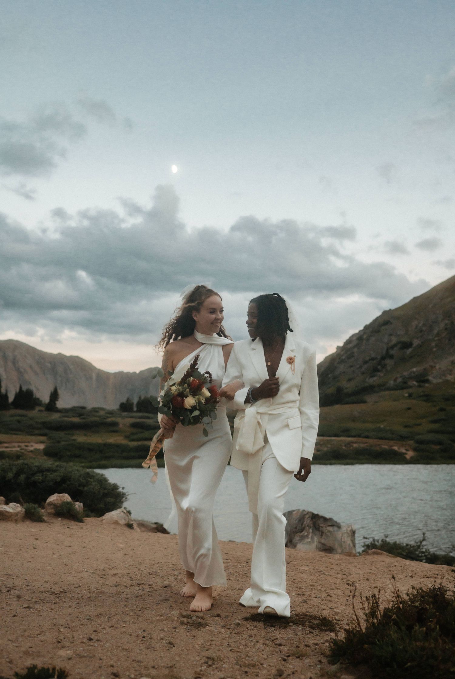 Tiny wedding ceremony unfolds along a dramatic mountain lake shoreline with the sky reflecting in the water.