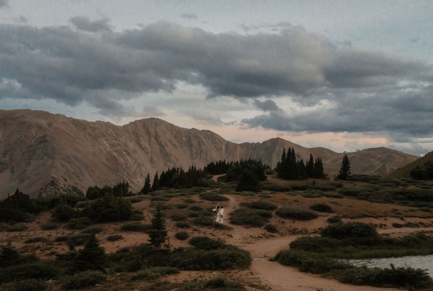 Winding dirt path through rugged mountain landscape with dramatic cloudy sky at dusk.