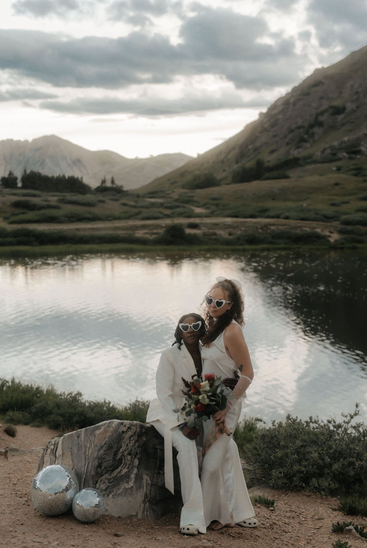 A romantic photoshoot by a serene mountain lake at sunset shows a couple in white outfits sitting on rocks.