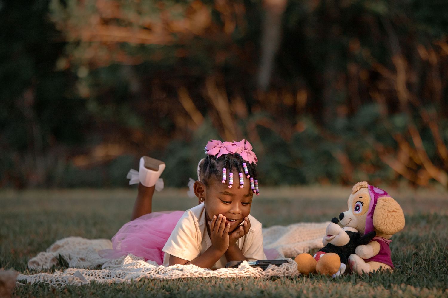 Child laying in grass at sunset with stuffed animal toys and wearing pink dress and floral headband.