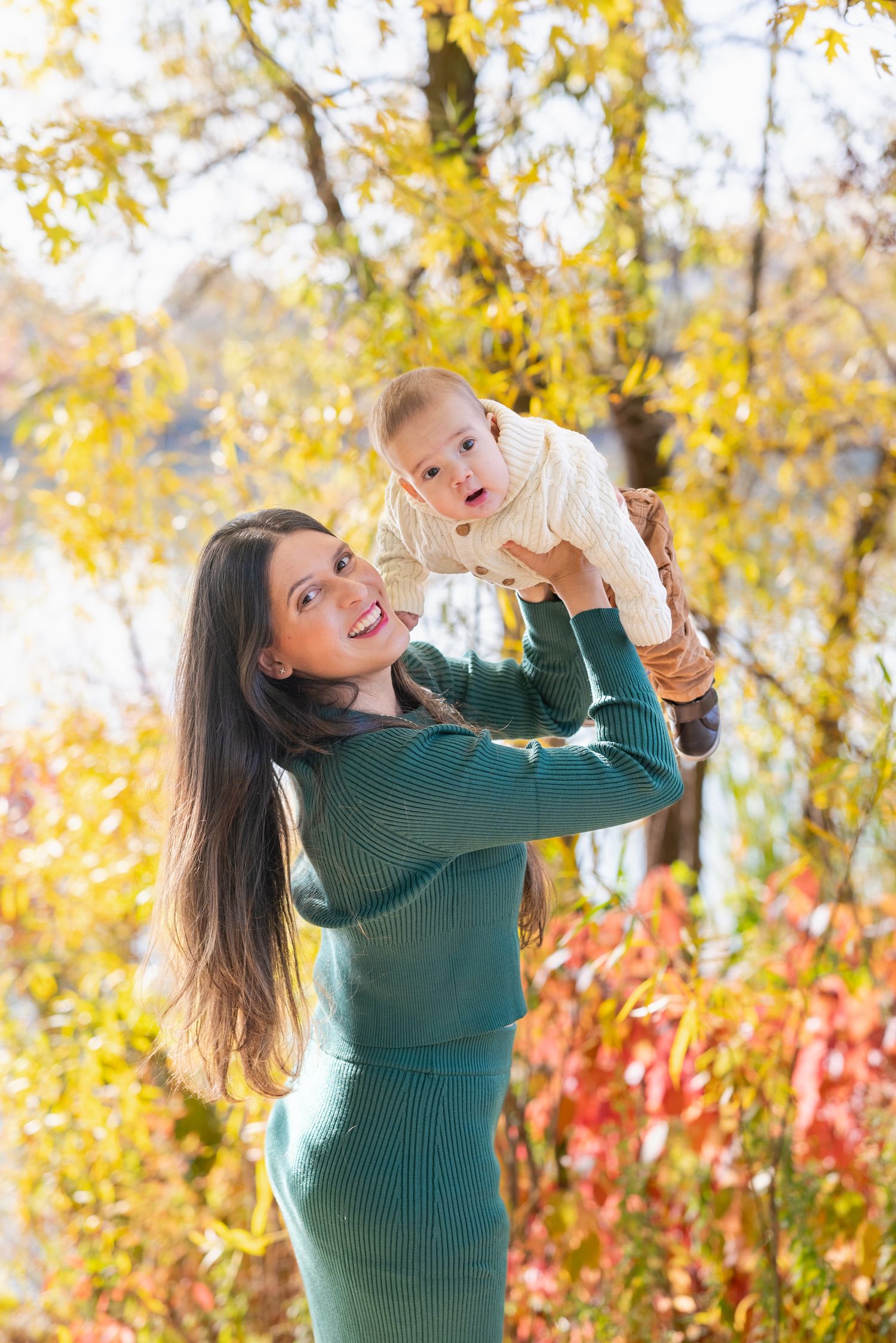 Mother in green sweater plays with baby against autumn foliage backdrop.