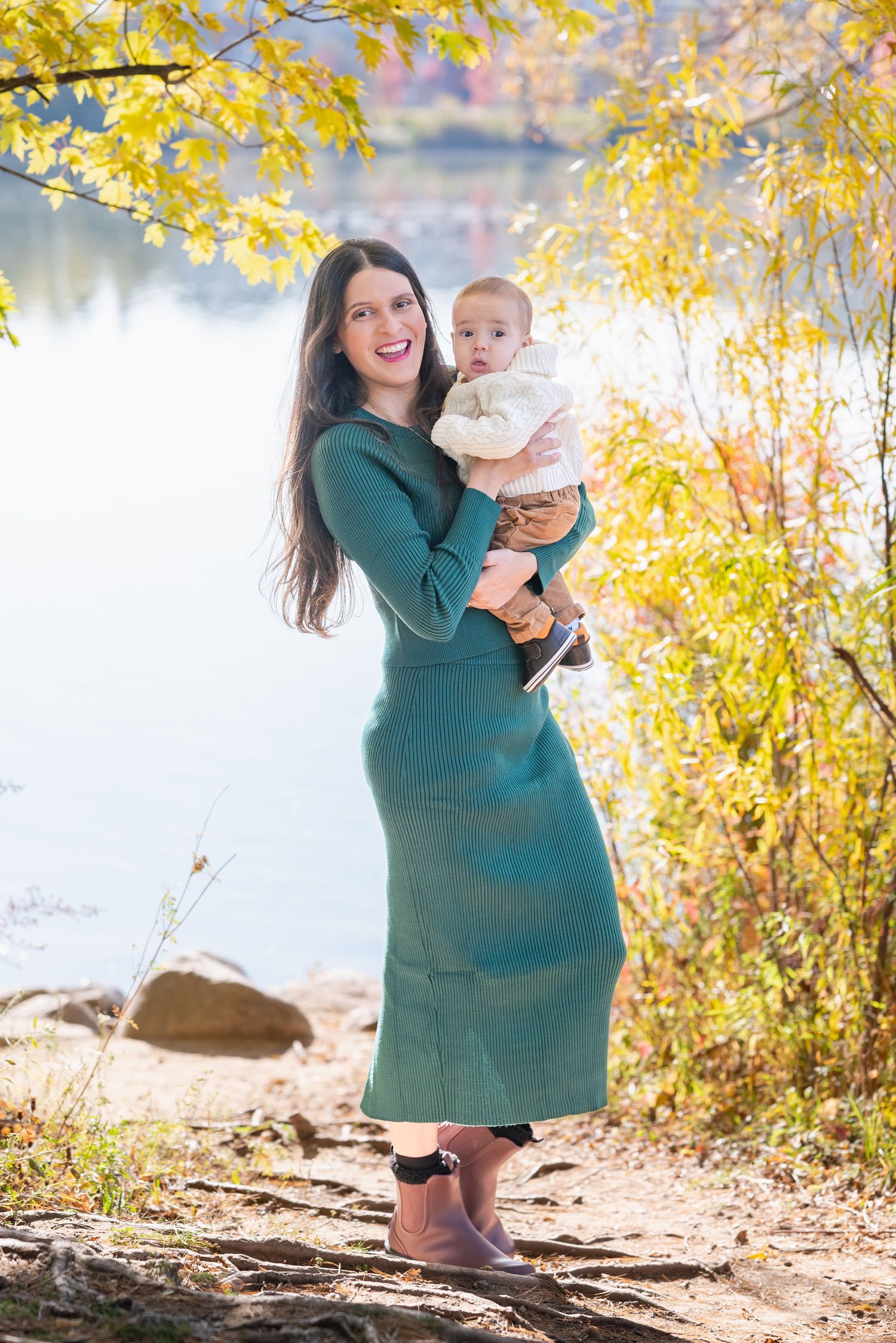 Mother in teal dress plays with baby by lakeside during fall season.