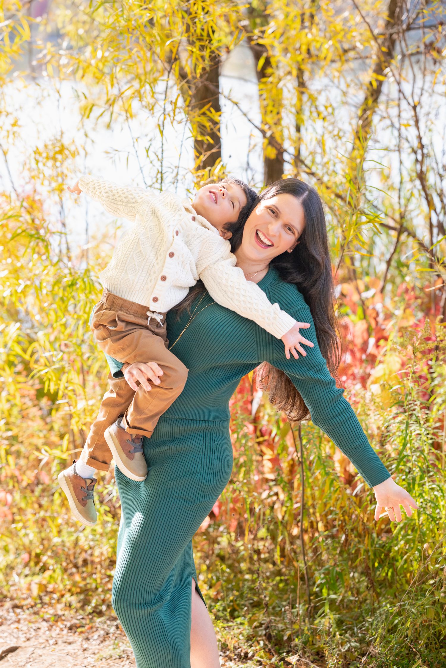 Woman in green dress playfully holds baby during autumn outdoor photoshoot.