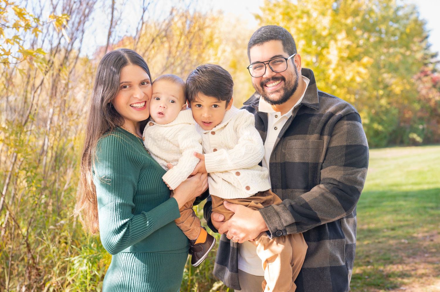 Family poses outdoors in casual attire during fall season with golden leaves in background.