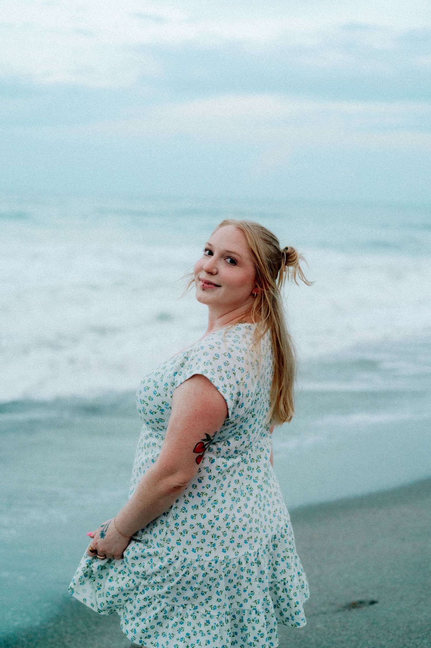 Woman in floral dress twirls on a misty beach with waves and cloudy turquoise sky in the background.
