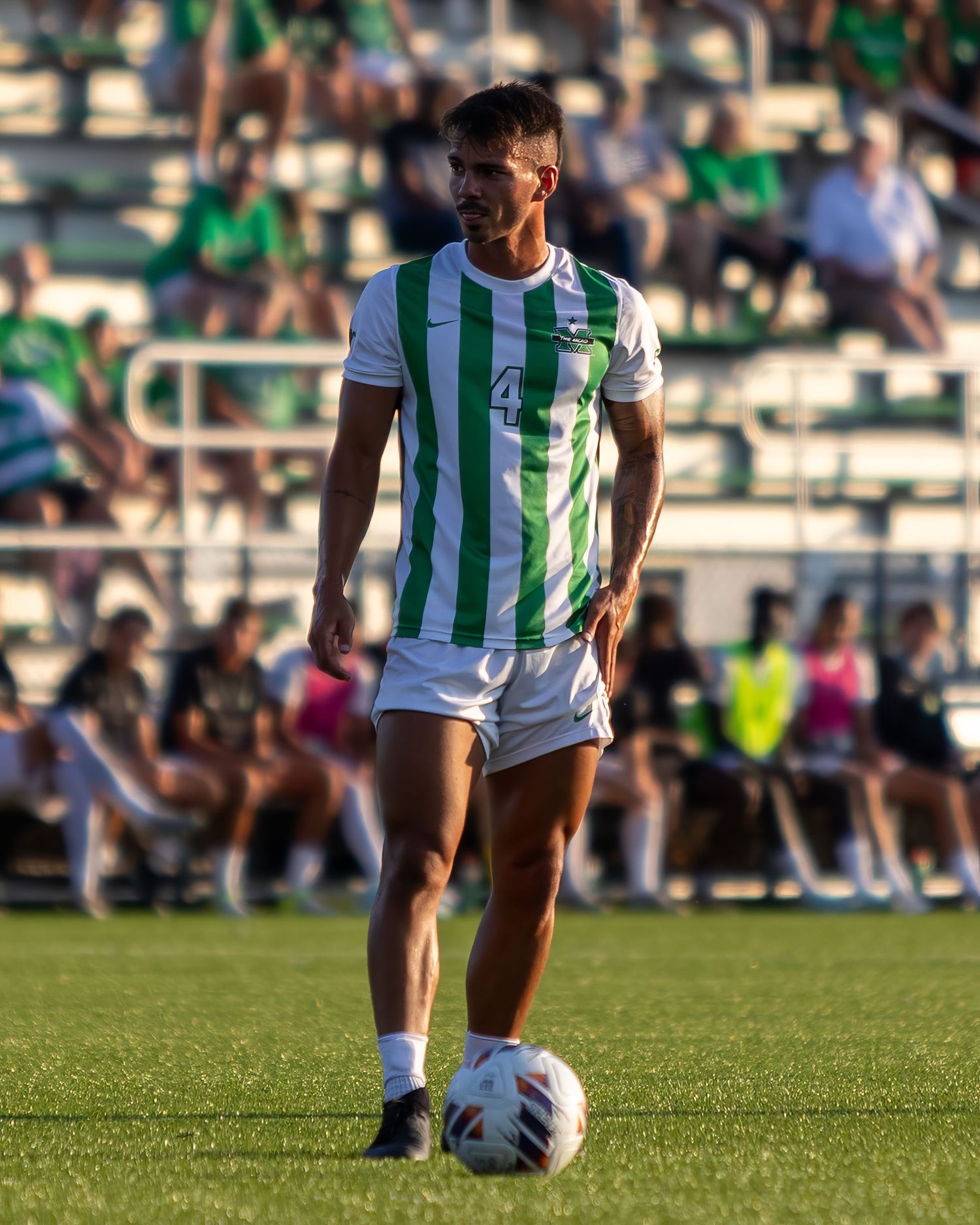 A soccer player in green and white striped jersey stands with ball on a sunny grass field during a match.