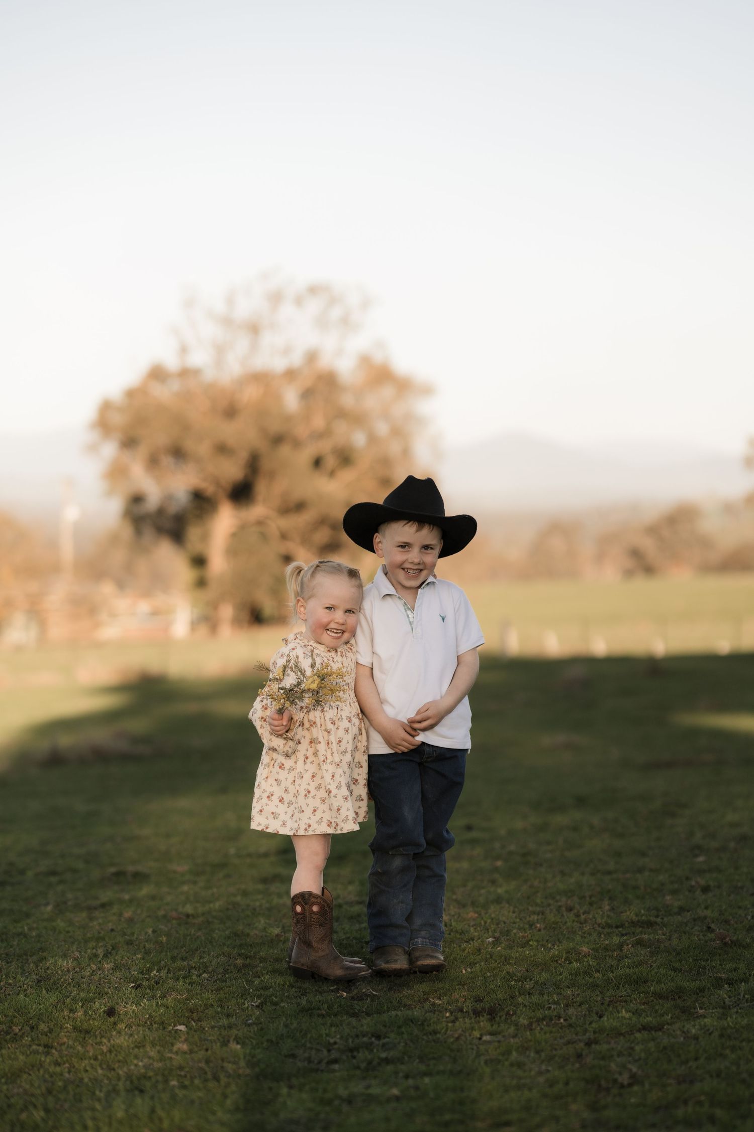 Two children holding hands while walking together on a grassy hill at sunset.