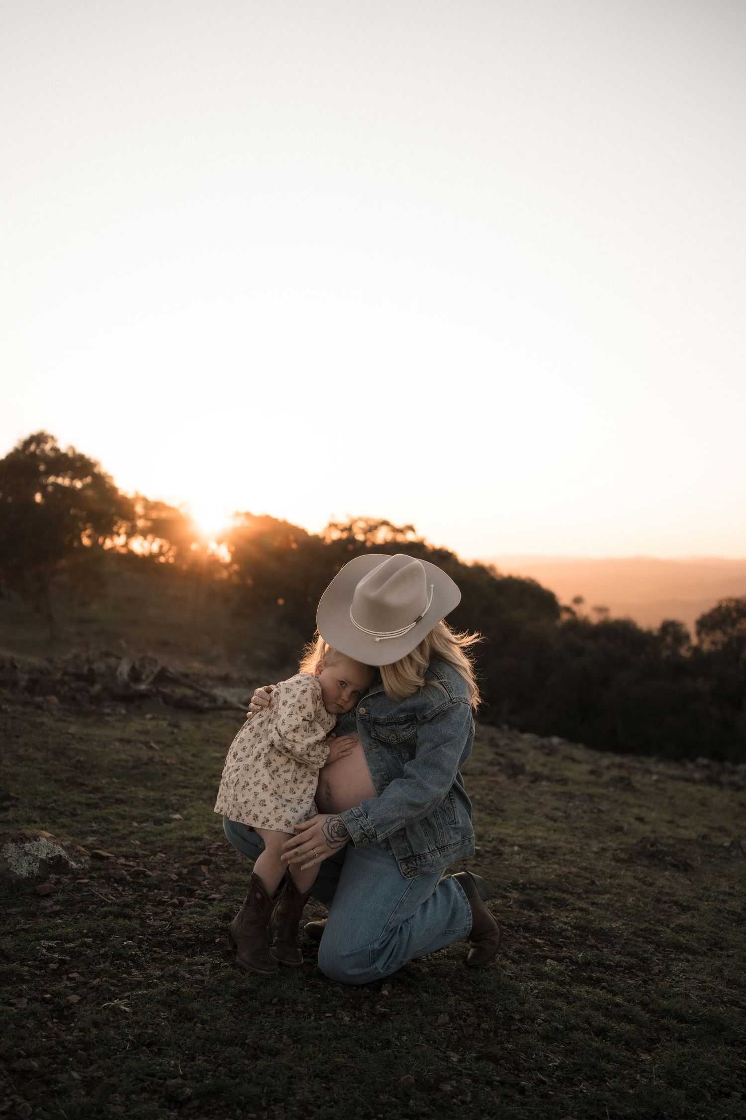 Two people share an intimate embrace at sunset on a grassy hilltop with golden sunlight filtering through trees.