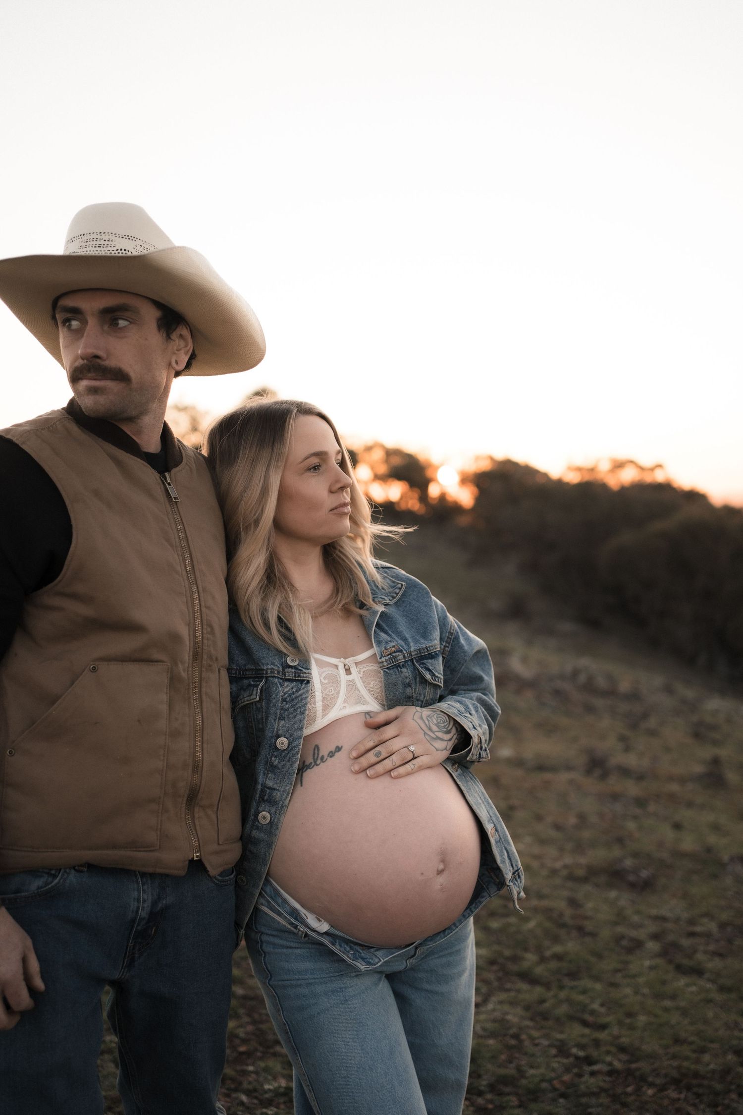 Maternity photos of couple in western wear standing together at sunset.