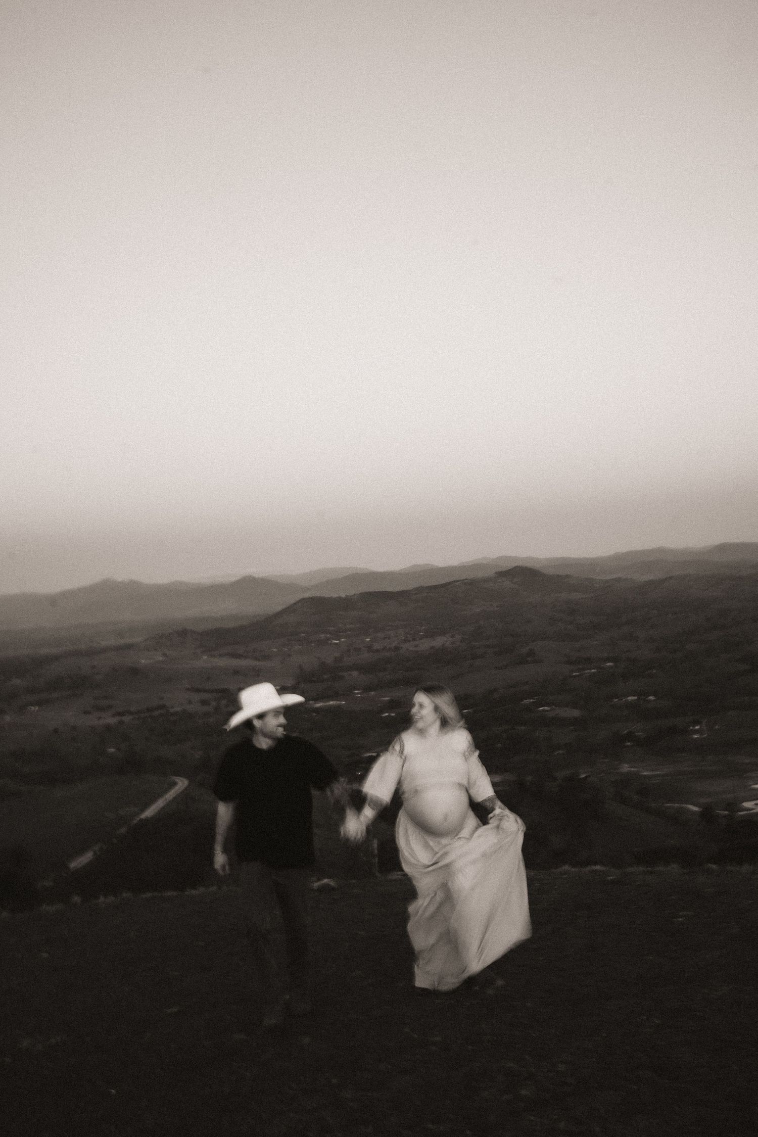 A black and white photo captures a couple on a hilltop overlooking a misty mountain landscape.