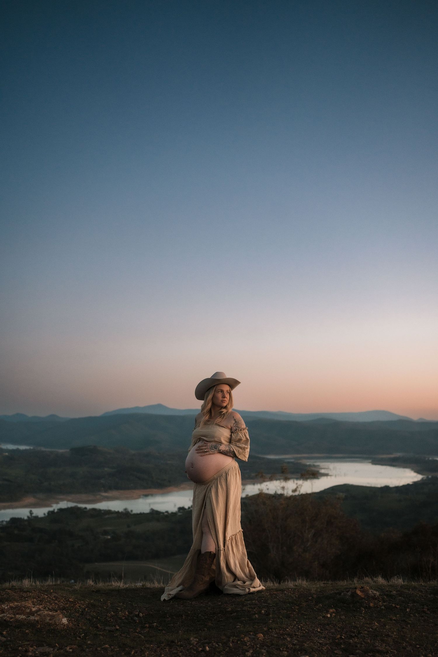 A silhouetted figure in flowing dress stands against a scenic mountain lake backdrop at dusk.