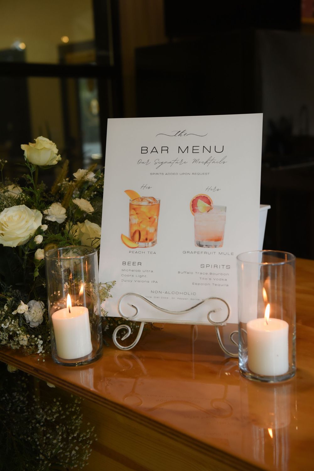 Wedding bar menu and white candles displayed on an elegant wooden surface with floral accents.
