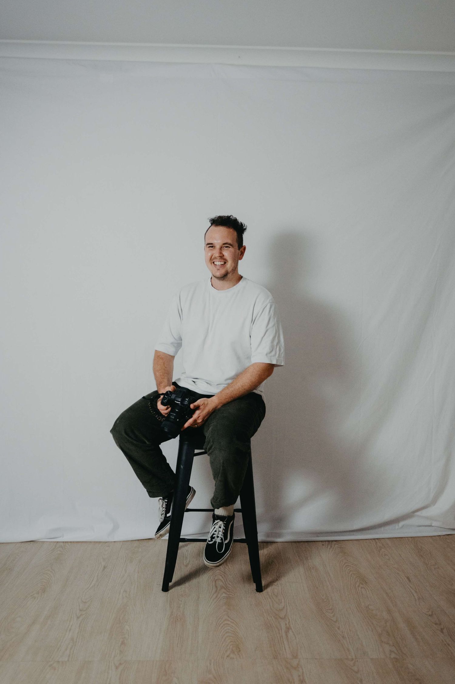 Minimalist portrait session against white backdrop featuring casual poses on black stool.