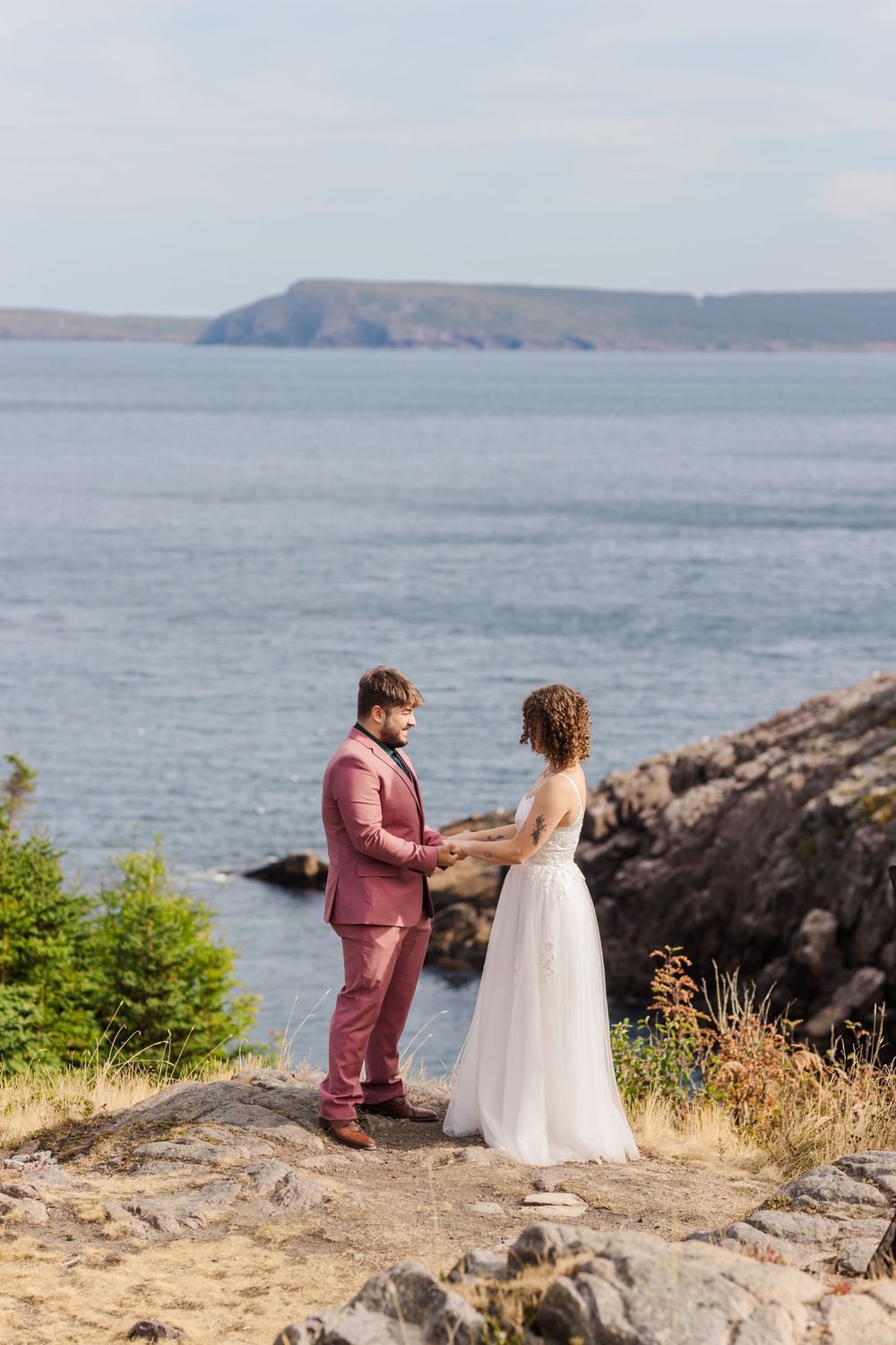 A couple shares a tender moment on a coastal cliff overlooking the ocean and rocky shoreline.
