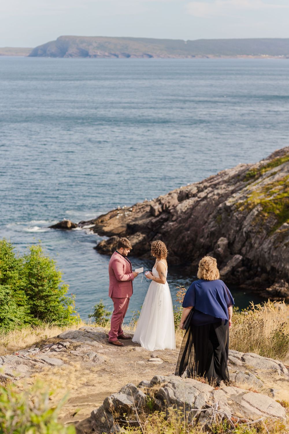 Wedding couple and photographers capture seaside photos on rocky coastal cliffs with ocean views.