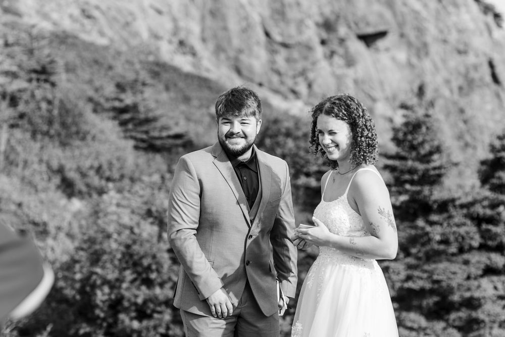 A happy couple in wedding attire share a joyful moment outdoors against a rocky cliff backdrop in black and white.