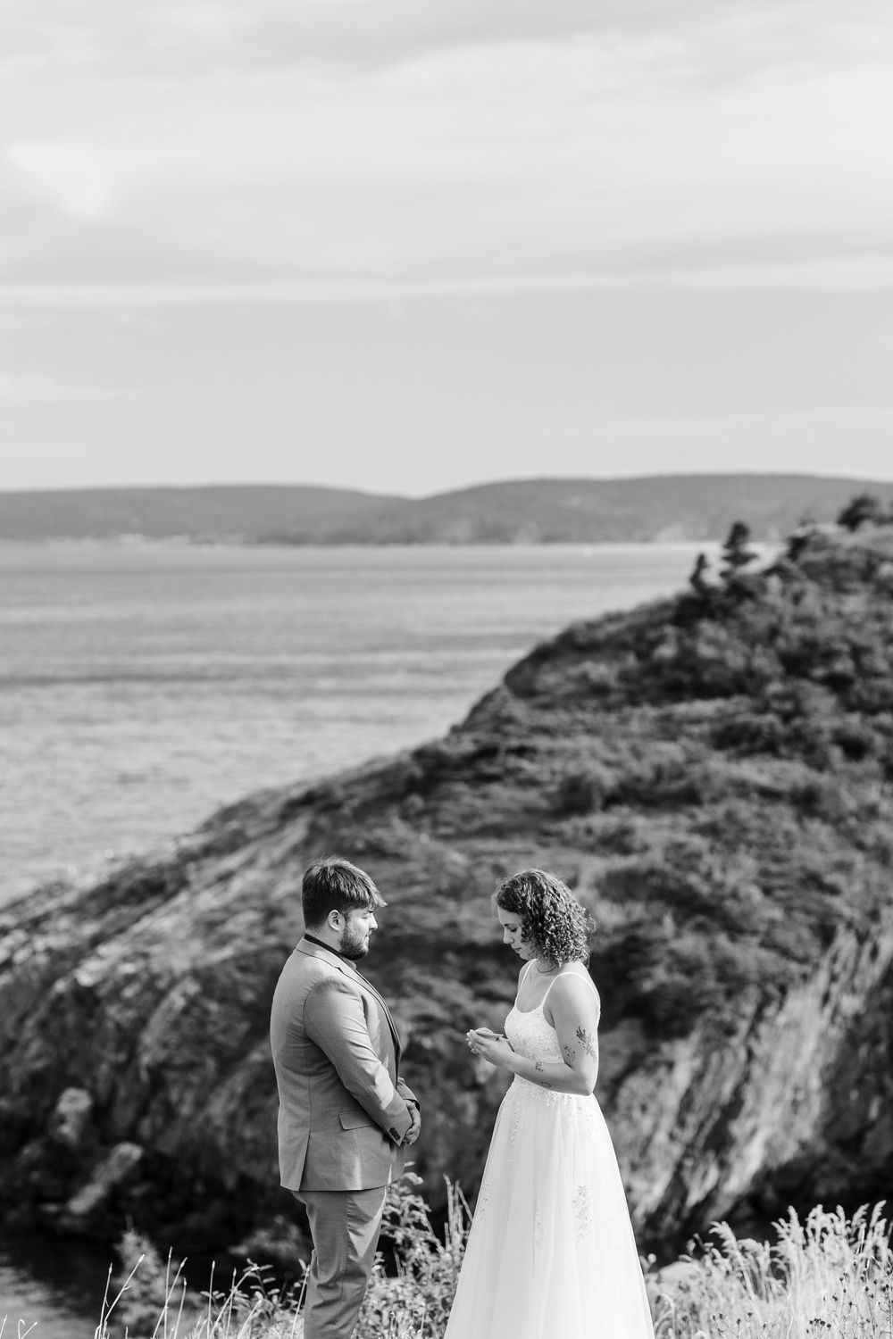 A couple shares a moment on a scenic coastal cliff overlooking the ocean in black and white.