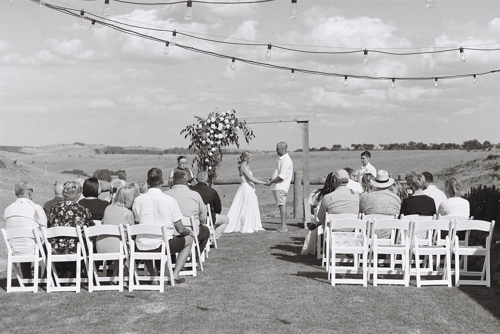 White chairs arranged for an outdoor wedding ceremony with string lights overhead against a cloudy sky.