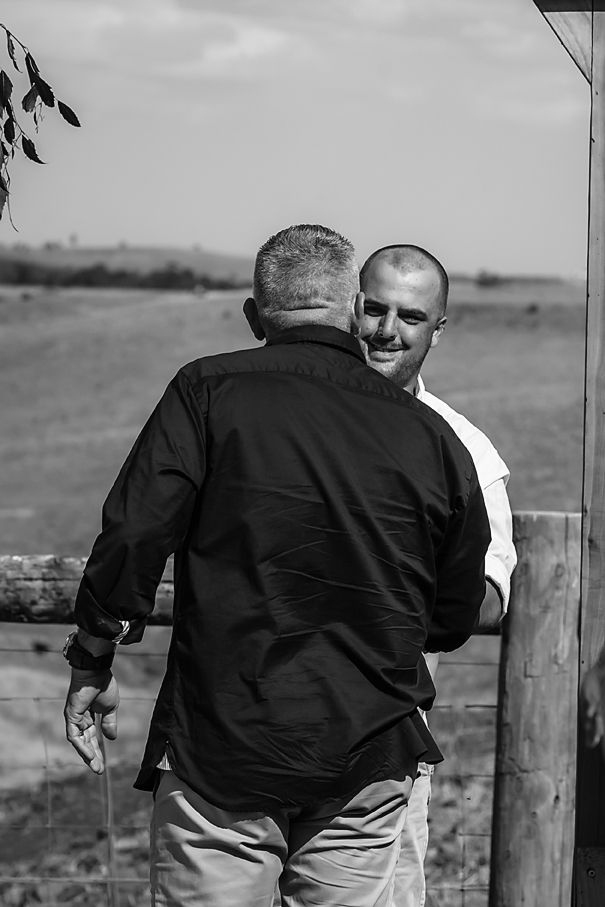Two people in black jackets stand at a wooden fence overlooking a rural landscape in black and white.
