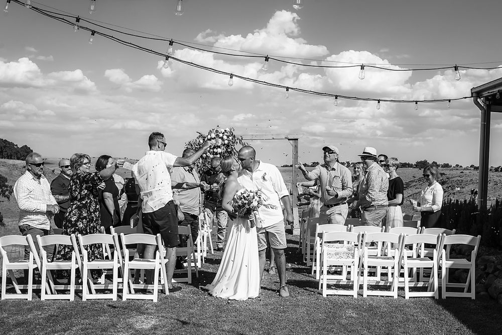 A black and white panoramic view of an outdoor wedding ceremony with rows of white chairs under string lights.