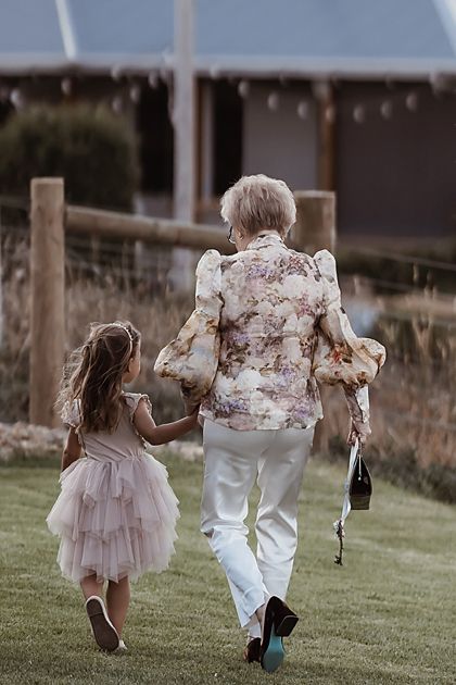 Two people walking hand in hand on grass, wearing soft pink and floral outfits in an evening setting.