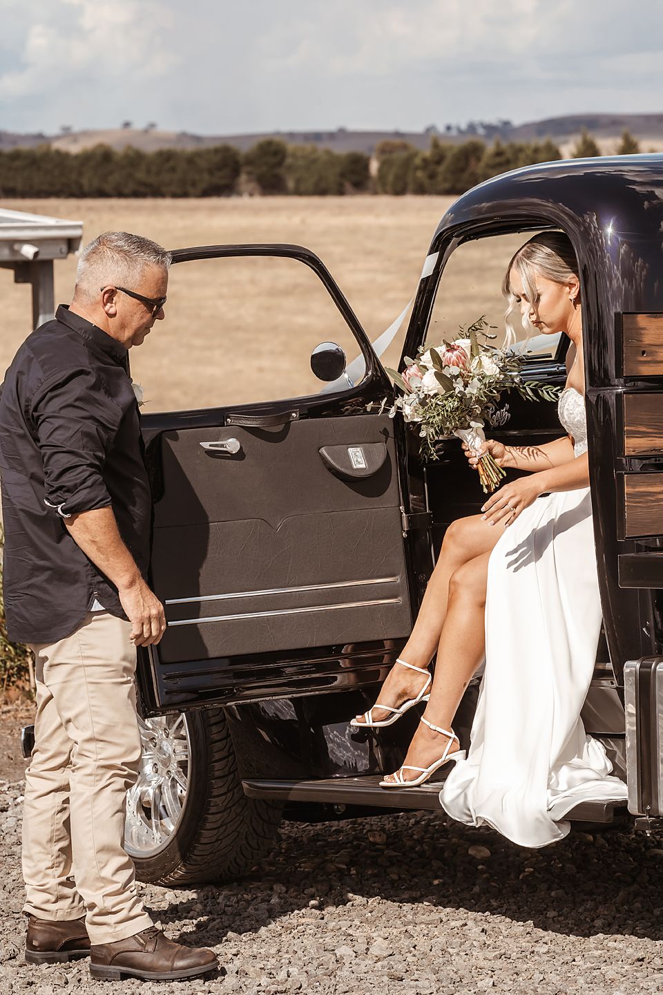 Wedding couple enters vintage black car with helper assisting bride in white dress.