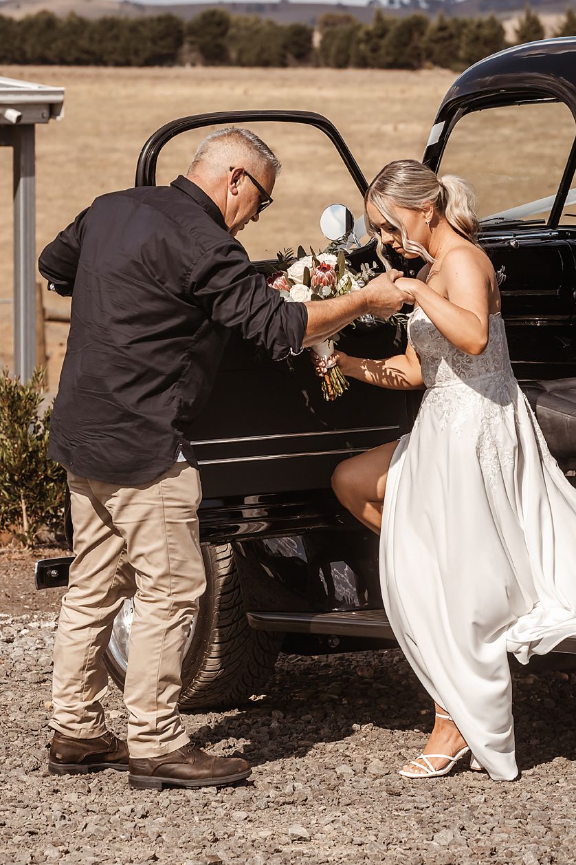 A wedding moment by a classic car where someone helps with final preparations outdoors.