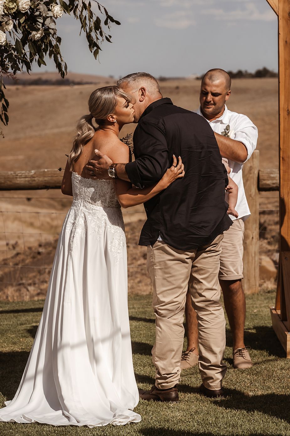 A wedding ceremony taking place outdoors with a rustic backdrop and floral arch.