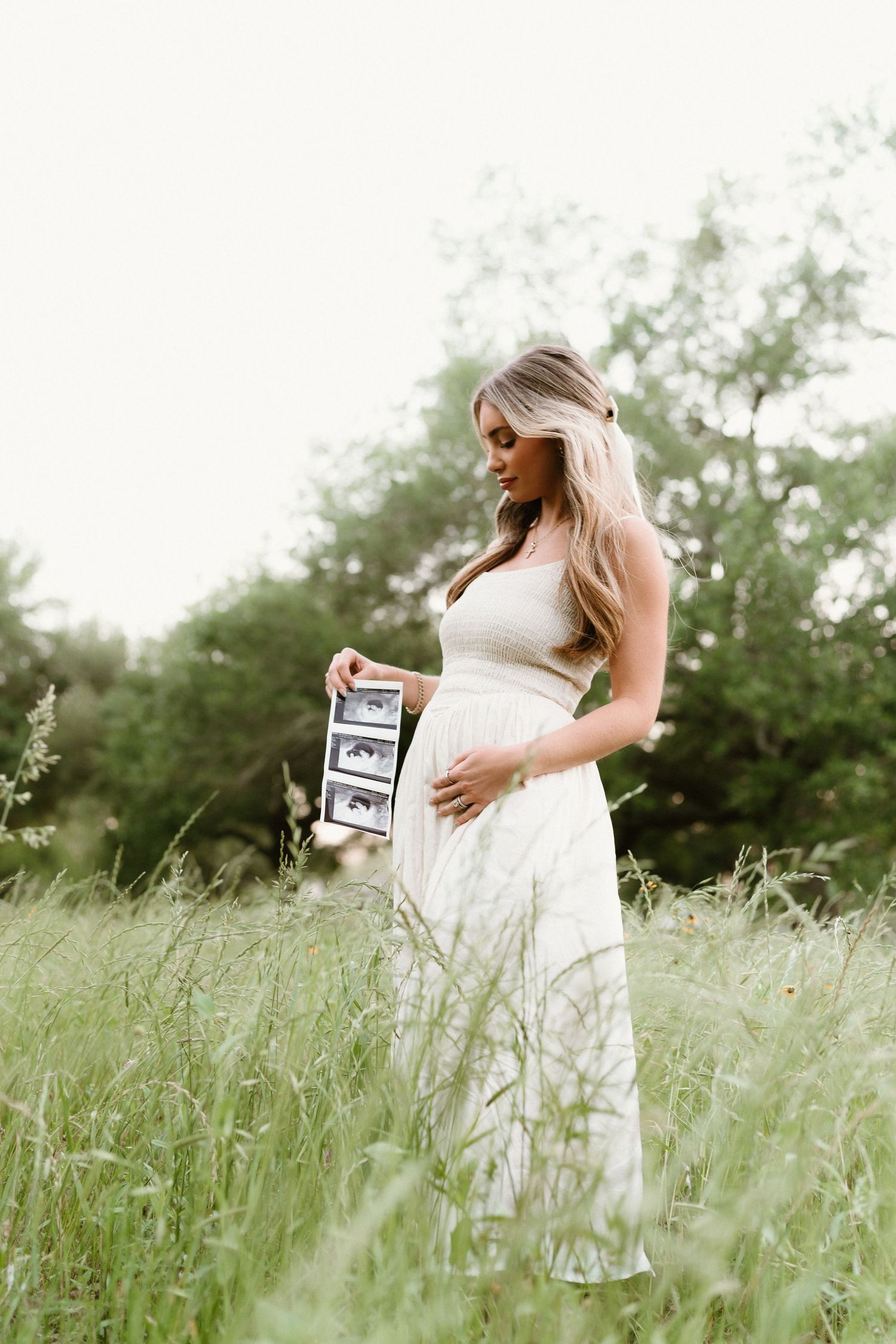 Woman in white dress holds chalkboard sign while standing in tall grass field.