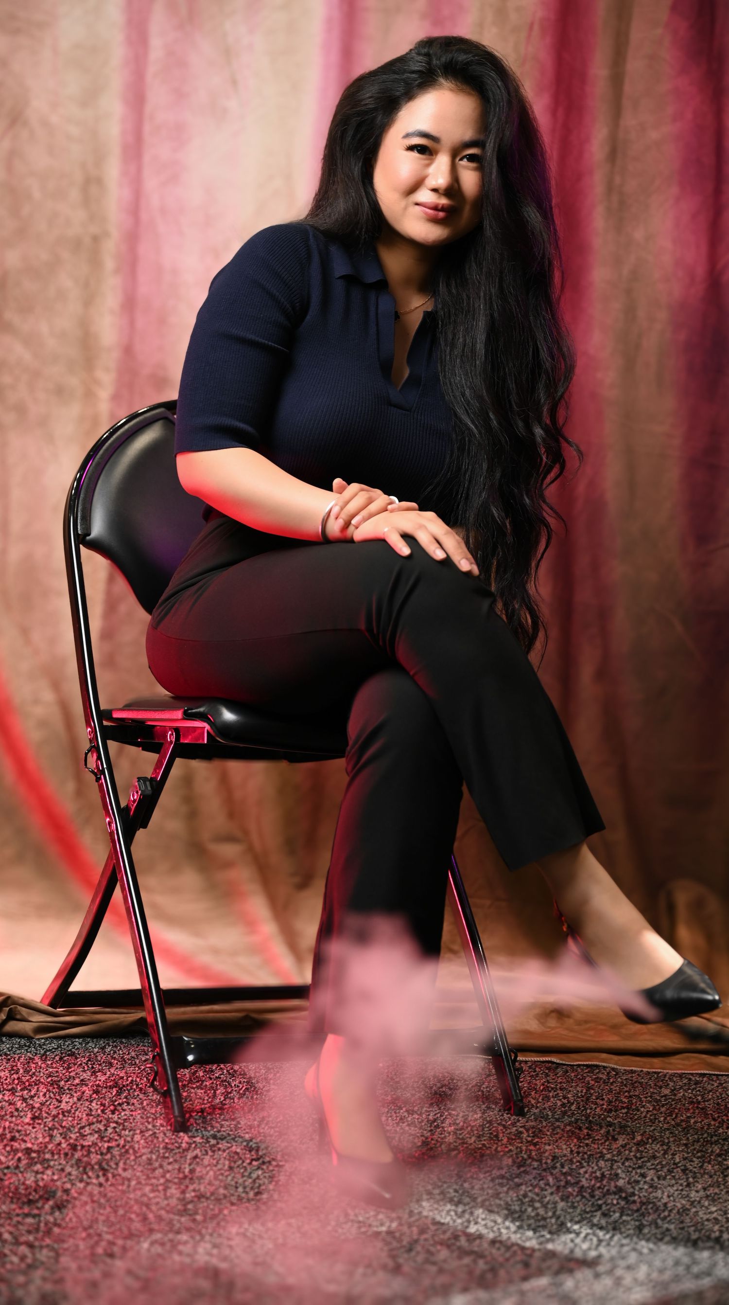 Person in black clothing poses on a chair against pink drapes on a carpeted floor in an artistic photoshoot.
