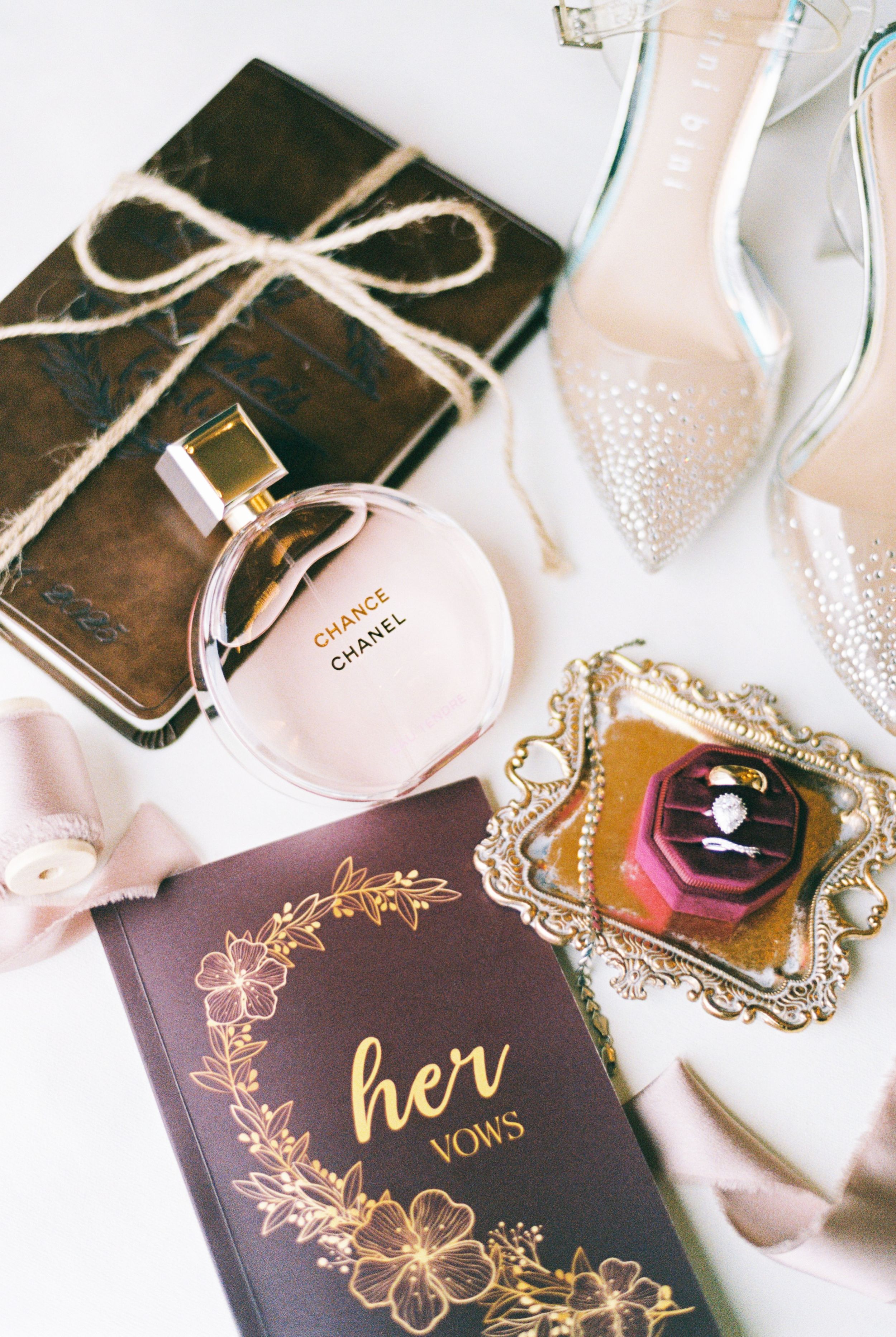 Wedding details including vow book, perfume, and accessories arranged artistically on white surface.