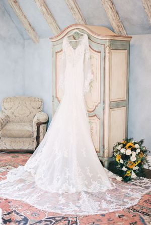 A white lace wedding dress with a dramatic train displayed on an ornate patterned carpet in an elegant room.
