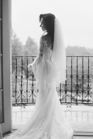 A bride in a flowing lace gown stands elegantly on a balcony with ornate iron railings in black and white photography.