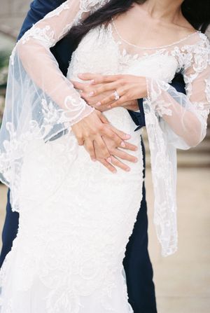 Close-up details of delicate lace and embroidery on a white wedding dress with hands visible.
