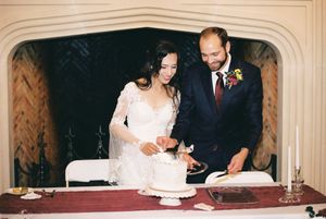 A couple in formal wedding attire cut their white wedding cake together at an elegant indoor reception.