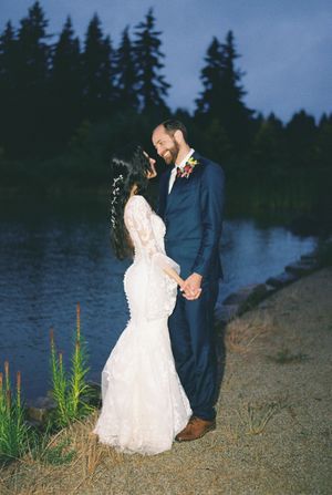 Couple in wedding attire sharing romantic moments at dusk by a serene lakeside with evergreen trees in the background.
