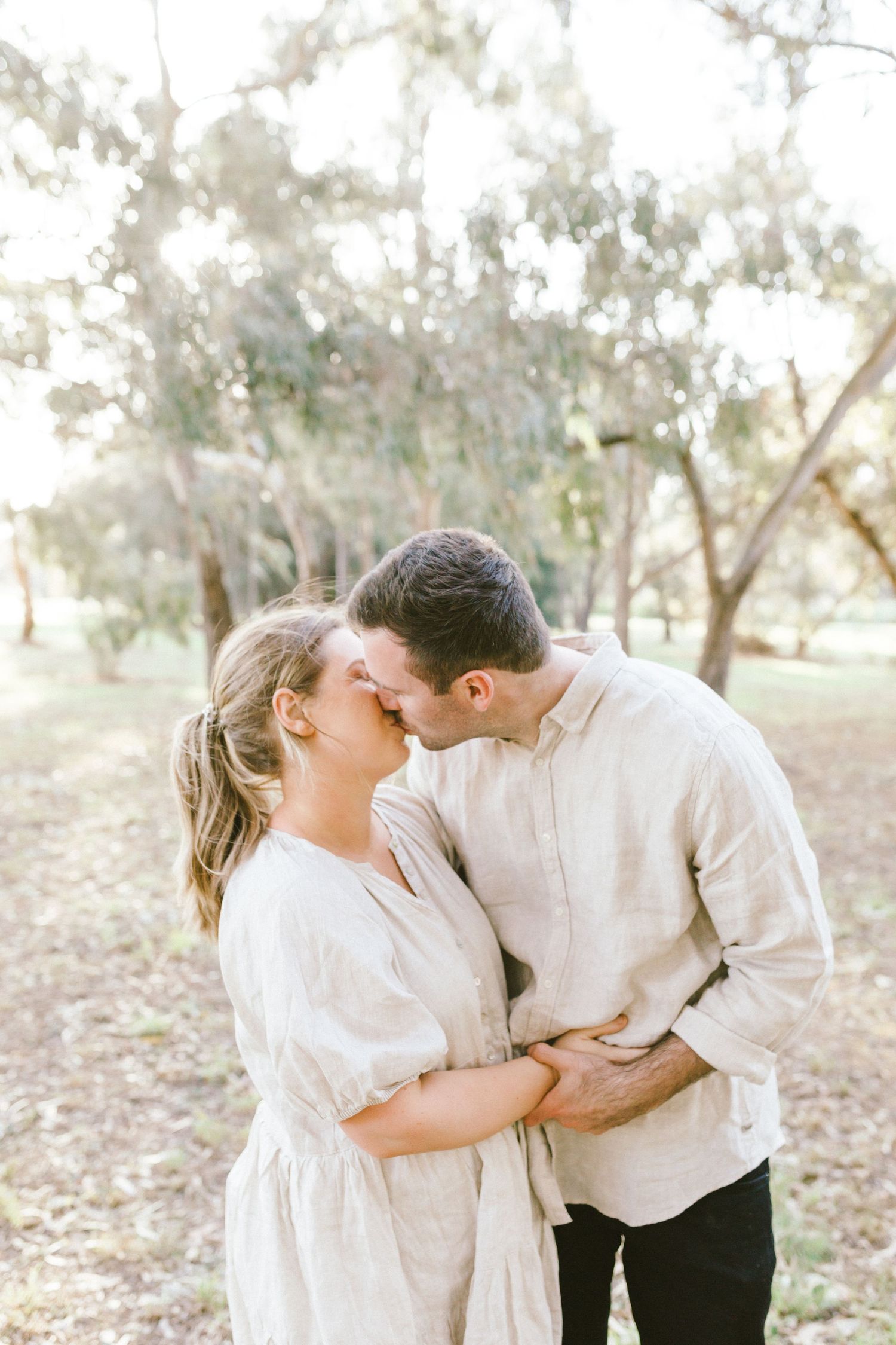 A romantic moment captured of a couple in white embracing outdoors in a sun-dappled wooded setting.