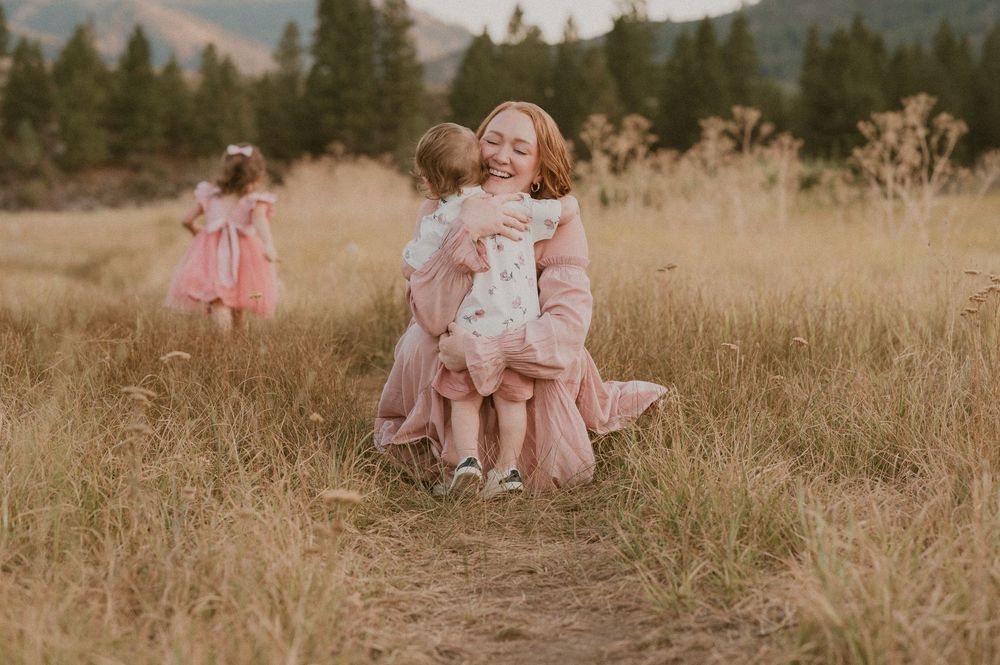 People in matching pink dresses gather in a golden field with mountains and pine trees in the background at sunset.