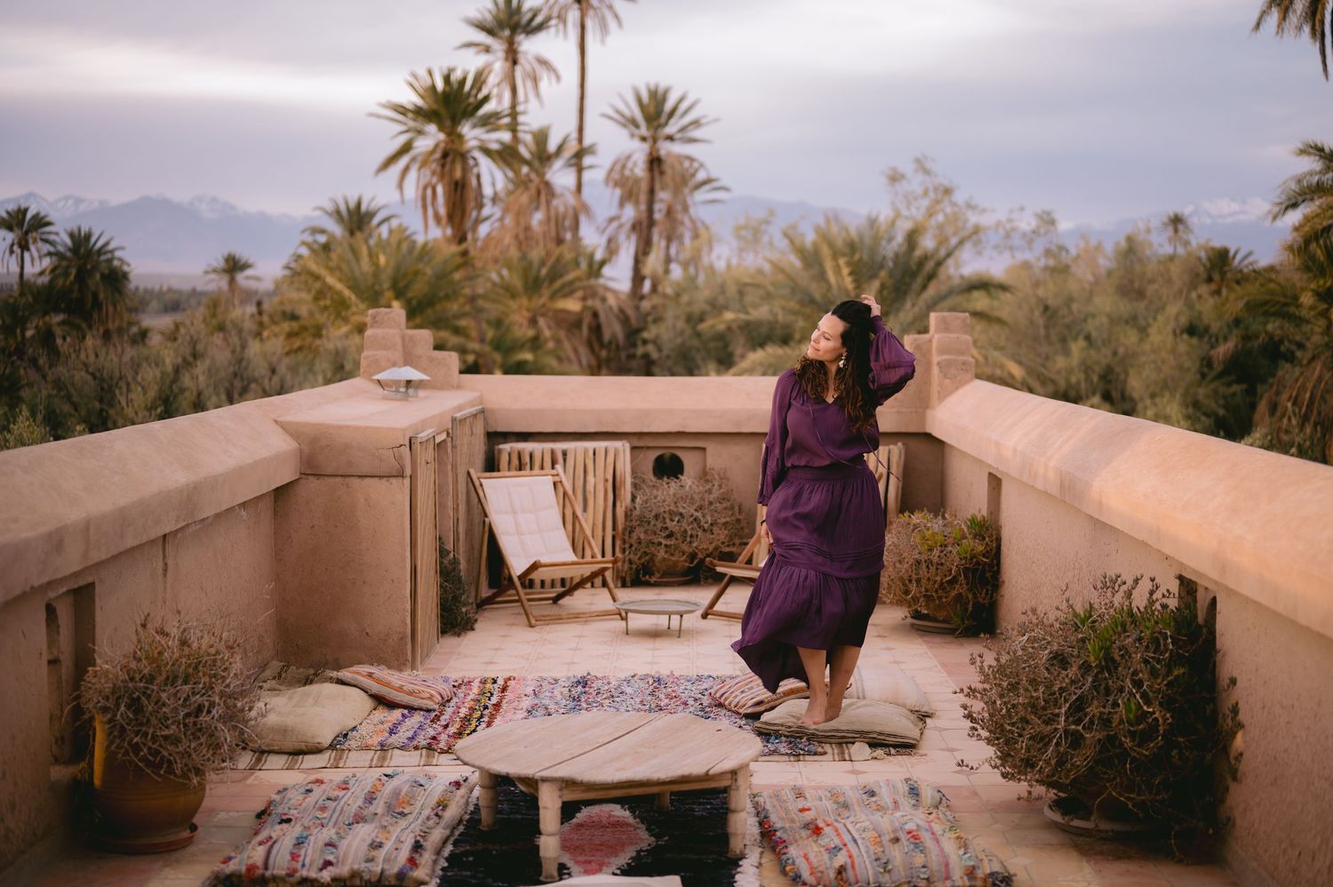 A rooftop terrace in Morocco decorated with rugs and cushions overlooking palm trees and mountains.