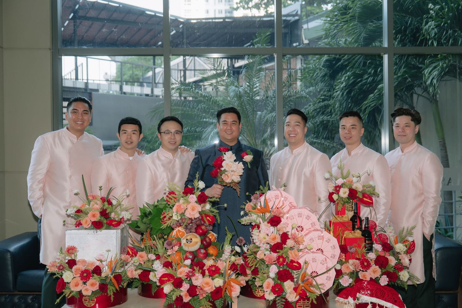 Groom with groomsmen in pink ao dai suits posing with red and pink floral arrangements at a Vietnamese wedding.