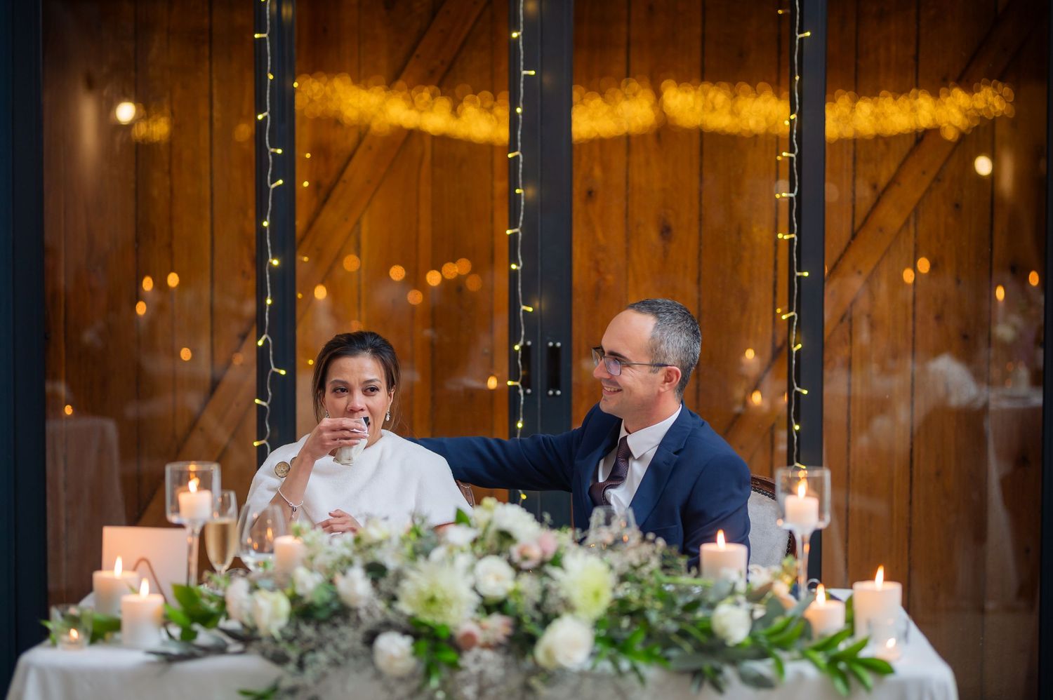 A rustic wooden barn wedding reception with twinkling string lights and elegant white floral centerpieces at long tables.
