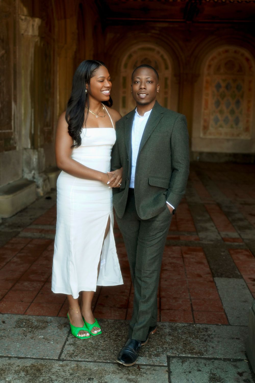A couple in formal attire poses for engagement photos at a historic venue with ornate stone architecture. Bethesda Terrace