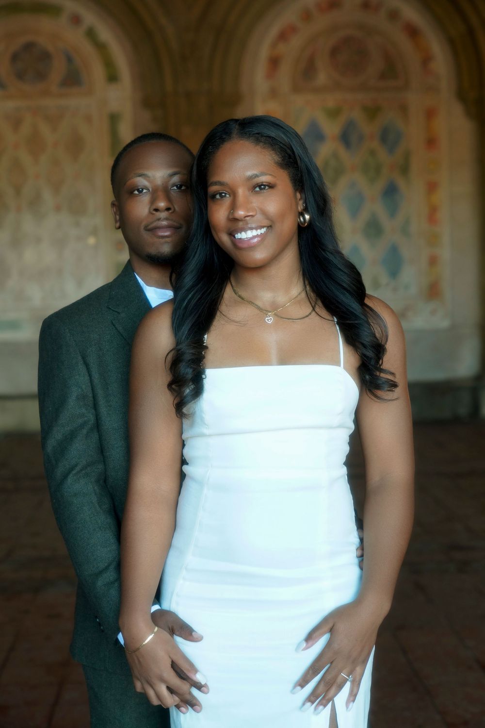 Couple stands together in formal attire against a decorative archway with diamond patterns in the background. Bethesda Terrace