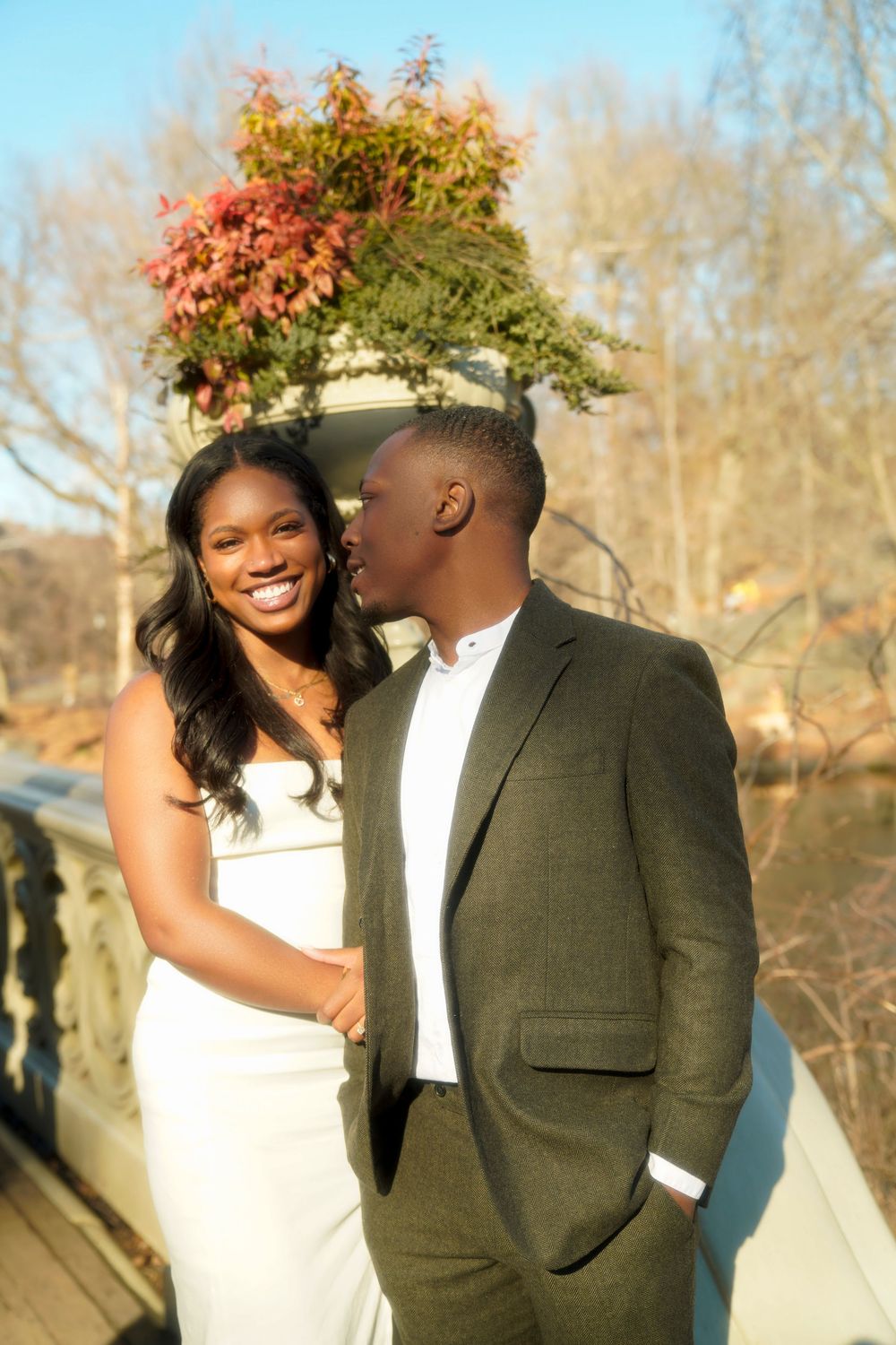 A couple shares a romantic moment together outdoors on a wooden deck during autumn with fall foliage in the background. central park