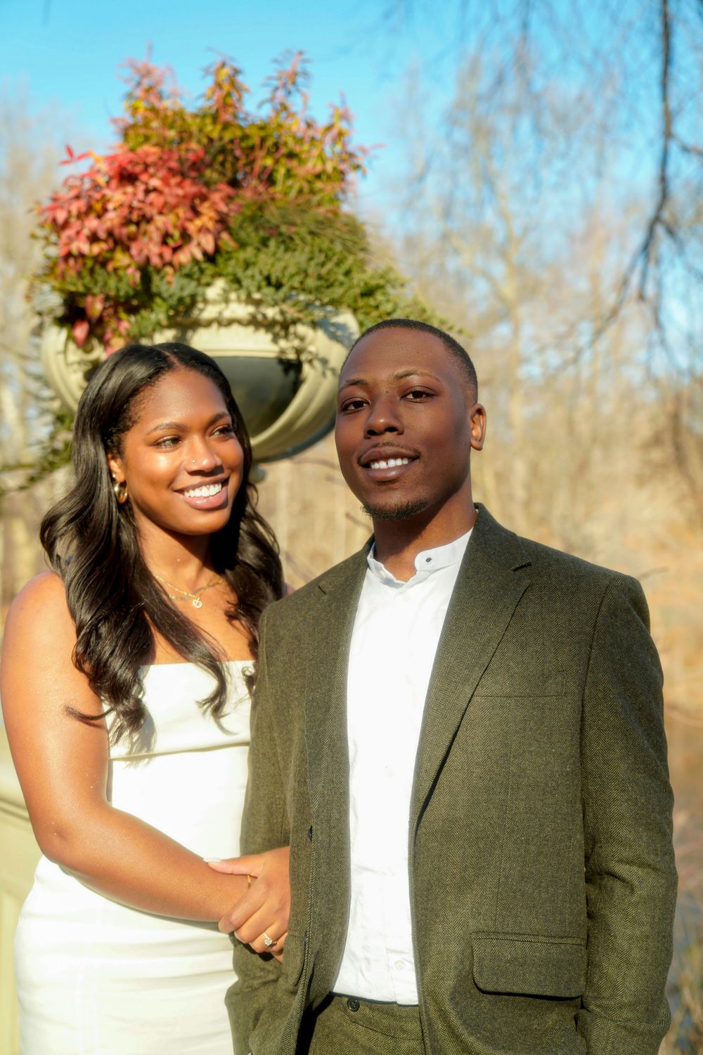 A happy couple in formal attire poses outdoors on a sunny day with fall foliage in the background. central park